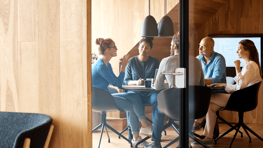 5 people sitting at a round conference table with glass door and media screen on the wall